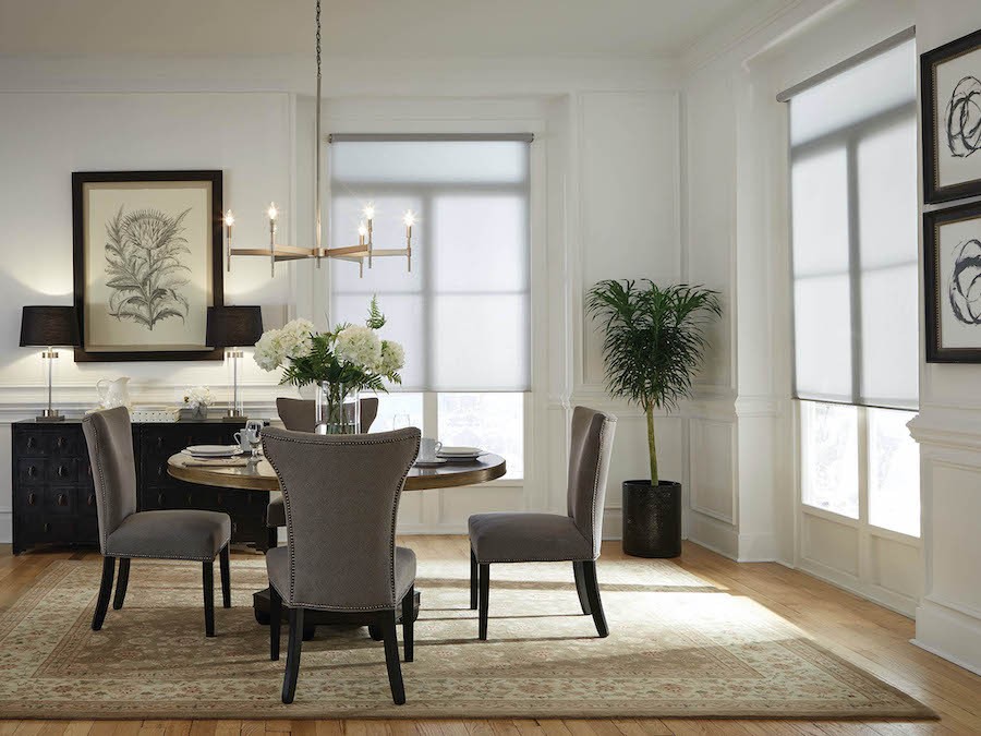 A corner dining room with chandelier and two large windows with motorized shades half drawn.
