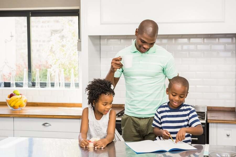 A father drinks a cup of coffee in a luxury kitchen while he helps his son and daughter with their homework.